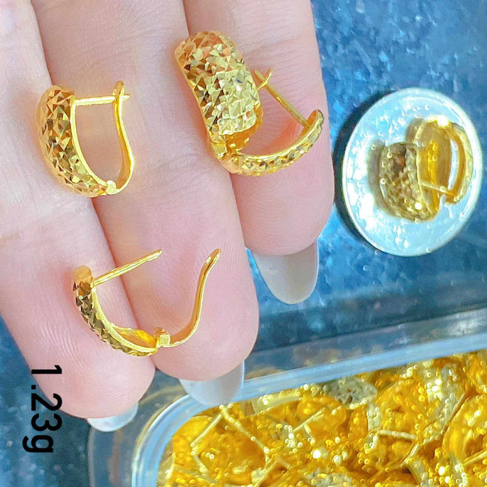 Gold hoop earrings held between fingers with a coin and container of gold pieces in the background.